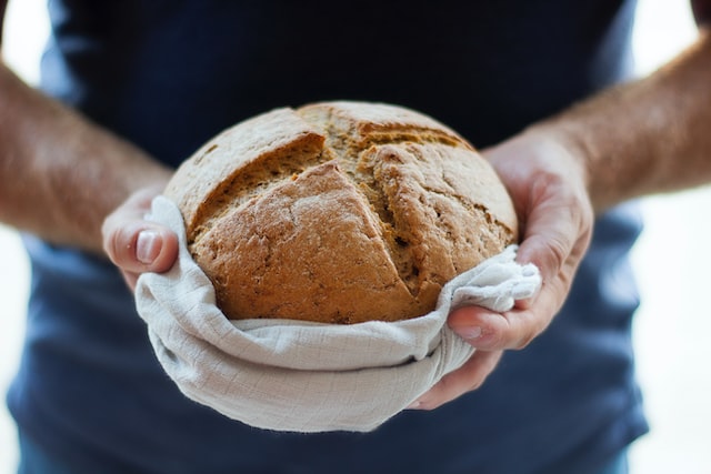 A man holds a baked loaf towards the camera, with a distinctive cross cut across the top.