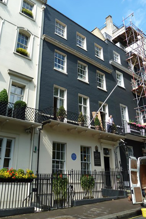 A Georgian townhouse. The ground floor is painted cream with three upper floors painted black. A man stands on the first-floor balcony.