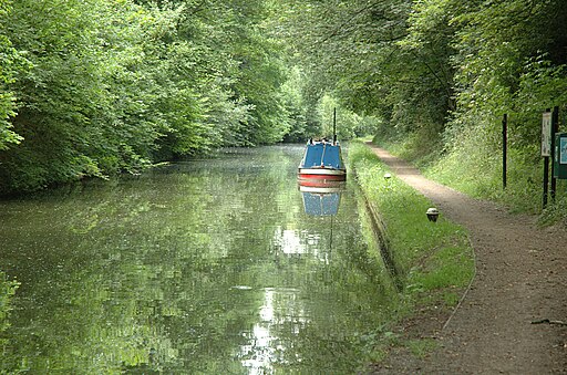 A canal with trees overhanging the lefthand side, and a tow path running along the righthand side. A canal boat is berthed against the bank.