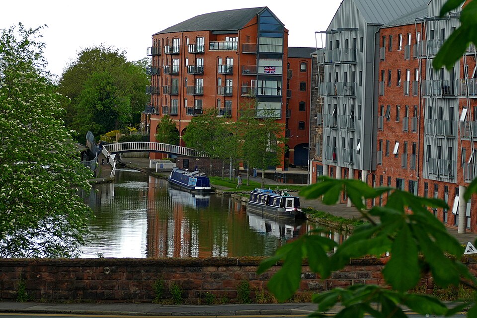 A canal flows beside a footpath, beyond which are apartment buildings in what looks like old industrial buildings. Two canal boats are berthed beside the tow path.